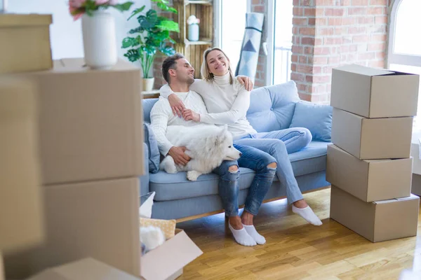 Young beautiful couple with dog kissing sitting on the sofa at new home around cardboard boxes