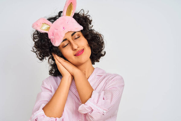 Arab woman with curly hair wearing pajama and sleep mask over isolated white background sleeping tired dreaming and posing with hands together while smiling with closed eyes.