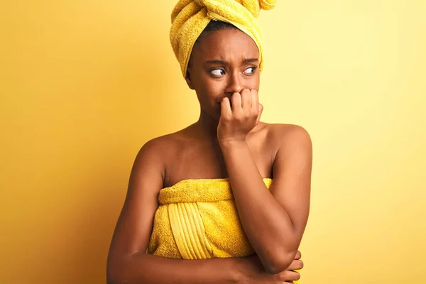 African american woman wearing towel after shower standing over isolated yellow background looking stressed and nervous with hands on mouth biting nails. Anxiety problem.