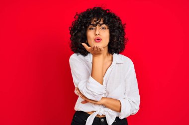 Kıvırcık saçlı, beyaz tişörtlü, izole edilmiş kırmızı arka planda genç bir Arap kadın havadaki güzel ve seksi elleriyle öpücük savururken kameraya bakıyor. Aşk ifadesi.