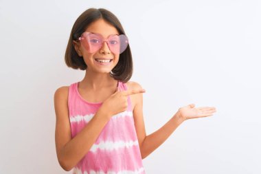 İzole edilmiş beyaz arka planda pembe tişört ve güneş gözlüğü takan genç ve güzel bir kız çocuğu kameraya hayret ediyor ve gülümsüyor..
