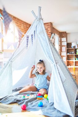 Genç, güzel sarışın kız anaokulunda oyuncaklarla eğleniyor. Evde Kızılderili çadırında oynarken mutlu gülümsüyor.