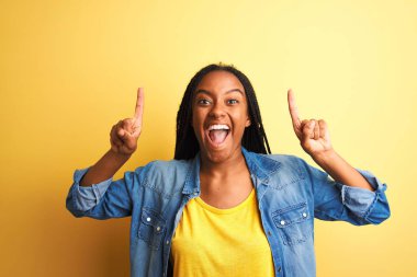 Genç Afro-Amerikalı kadın kot gömlek giyip izole edilmiş sarı arka planda dikilmiş şaşkın şaşkın gülüyor, parmaklarıyla işaret ediyor ve kollarını kaldırıyordu..