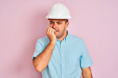 Güvenlik kaskı takan genç mimar pembe arka planda duruyor. Ağzı tırnak yiyen eller yüzünden gergin ve stresli görünüyor. Endişe sorunu.