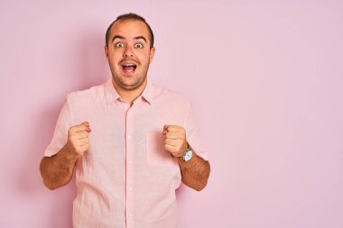 Soyutlanmış pembe arka planda duran zarif gömlekli genç adam kollarını kaldırıp açık gözlerle başarının şaşkınlığını ve şaşkınlığını kutluyor. Kazanan konsept.