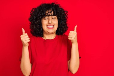 Kıvırcık saçlı genç Arap kadın izole edilmiş kırmızı arka plan üzerine günlük tişört giyiyor. Kolları havada ve gözleri kapalı zaferi kutlarken gülümsüyor. Kazanan konsept.