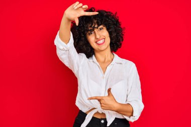 Kıvırcık saçlı, beyaz gömlekli, soyutlanmış kırmızı arka planda gülümseyen, elleri ve parmakları mutlu yüzlü genç bir Arap kadın. Yaratıcılık ve fotoğrafçılık kavramı.