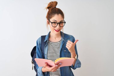 Sırt çantası takan kızıl saçlı kız izole edilmiş beyaz arka plan üzerine kitap okuyor ve gülümseyen yüzüyle baş parmağıyla işaret ediyor.