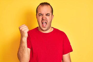İzole edilmiş sarı arka planda dikilen kırmızı tişörtlü genç adam öfkeyle bağırıyor, deliriyor ve el kaldırarak bağırıyor. Öfke kavramı.
