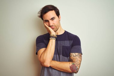 İzole edilmiş beyaz arka plan üzerine günlük tişört giyen yakışıklı genç adam kollarını kavuşturup depresyondan sıkılmış ve yorgun görünüyor..