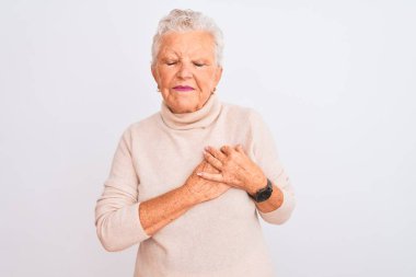Üst düzey, gri saçlı, balıkçı yaka kazak giyen, izole edilmiş beyaz arka plan üzerinde duran, elleri göğsünde, gözleri kapalı ve yüzünde minnet dolu bir jestle gülümseyen kadın. Sağlık kavramı.