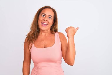 Orta yaşlı olgun bir kadın beyaz soyutlanmış arka planda durup gülümsüyor ve başparmağıyla yan tarafa bakıyor..