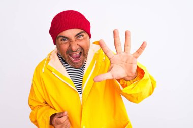 İzole edilmiş beyaz arka planda duran, yağmurluk ve yün şapka giyen orta yaşlı bir adam beş numarayı gösterirken kendinden emin ve mutlu bir şekilde gülümsüyor..