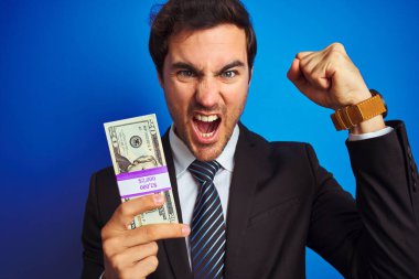 Takım elbiseli genç ve yakışıklı bir işadamı mavi arka planda dolarları elinde tutarken sinirli ve sinirli bir şekilde bağırıyor, çıldırıyor ve bağırıyor.