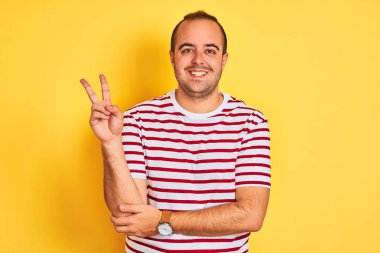 Sıradan çizgili tişört giyen genç adam izole edilmiş sarı arka planda dikiliyor ve kameraya gülümsüyor zafer işareti yapıyor. İki numara..