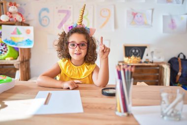 Gözlüklü ve tek boynuzlu at diademi takmış güzel bir çocuk anaokulundaki masada oturmuş başarılı bir fikirle işaret ediyor. Heyecanlı ve mutlu. Bir numara..