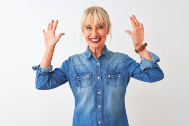 İzole edilmiş beyaz arka plan üzerinde duran sıradan bir kot gömlek giyen orta yaşlı kadın on numaralı parmaklarıyla işaret ederken kendinden emin ve mutlu bir şekilde gülümsüyor..