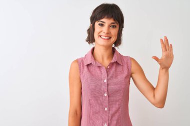 İzole edilmiş beyaz arka planda duran kırmızı tişörtlü genç ve güzel bir kadın beşinci parmaklarıyla işaret ederken kendinden emin ve mutlu bir şekilde gülümsüyor..