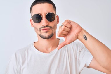 Güneş gözlüğü takan genç yakışıklı bir adam ve izole edilmiş arka planda, kızgın yüzlü, negatif işaretli, baş parmakları aşağıda, reddedilme kavramından hoşlanmadığını gösteren bir günlük tişört giyiyor.