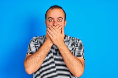 İzole edilmiş mavi arka planda duran çizgili donanma tişörtü giyen genç adam yanlışlıkla ağızlarını kapatarak şok geçirdi. Gizli konsept.