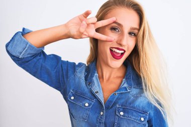 İzole edilmiş beyaz arka plan üzerinde duran sıradan bir kot gömlek giyen genç ve güzel bir kadın. Yüzünde parmakları olan barış sembolü yapıyor. Gülümsüyor, zaferini gösteriyor.