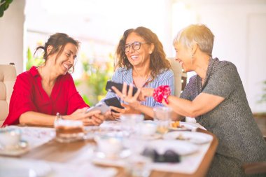 Orta yaşlı kadınlarla buluşup, öğle yemeği yiyip, kahve içiyorlar. Olgun arkadaşlar mutlu bir şekilde gülümsüyor. Güneşli bir günde evde akıllı telefon kullanıyorlar.