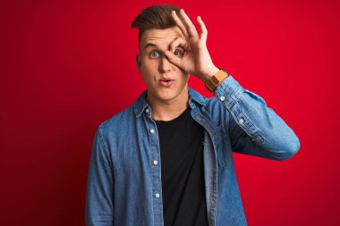 İzole edilmiş kırmızı arka plan üzerinde duran kot gömlekli genç yakışıklı adam şaşırmış yüz ifadesiyle şaşırmış, parmaklarının arasından bakan bir hareket yapıyor. İnançsız ifade.
