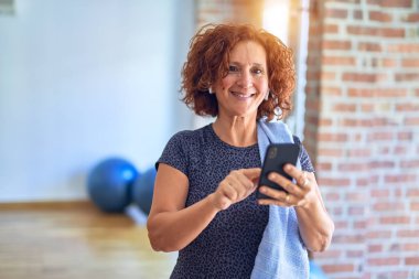Orta yaşlı güzel sporcu kadın gülümsüyor. Yüzünde gülümsemeyle spor salonunda akıllı telefon ve kulaklıkla dikiliyor.