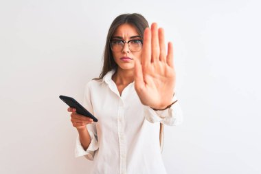 Gözlük takan güzel bir iş kadını izole edilmiş beyaz arka plan üzerinde akıllı telefon kullanıyor. Açık elle dur işareti yapıyor. Ciddi ve kendinden emin bir ifadeyle, savunma jesti.
