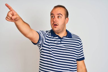 İzole edilmiş beyaz arka plan üzerinde duran sıradan çizgili polo giyen genç adam parmağıyla işaret ederek ileriyi işaret ediyor, ağzı açık şaşkın bir ifade, ön tarafta bir şey.