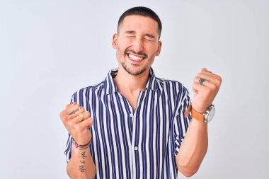 Deniz çizgili gömlek giyen genç yakışıklı adam izole edilmiş arka planda mutlu ve heyecanlı bir şekilde kollarını kaldırıp, gülümseyerek ve başarı için çığlık atarak kazanan jest yapıyor. Kutlama konsepti.