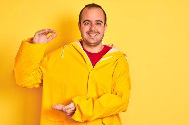 Yağmurluk giyen genç adam izole edilmiş sarı arka planda duruyor. Ellerinde büyük ve büyük boy işareti var. Ölçü sembolü. Kameraya bakıp gülümsüyor. Ölçüm konsepti.