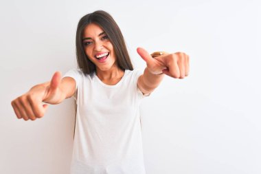 Young beautiful woman wearing casual t-shirt standing over isolated white background approving doing positive gesture with hand, thumbs up smiling and happy for success. Winner gesture.
