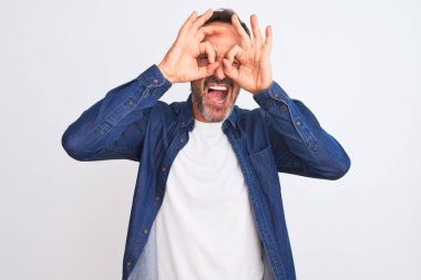 Mavi kot gömlek giyen orta yaşlı yakışıklı bir adam izole edilmiş beyaz arka planda dikiliyor dürbün gibi jest yapıyor, dili dışarı çıkarıyor, gözleri parmaklarından bakıyor. Çılgın ifade.