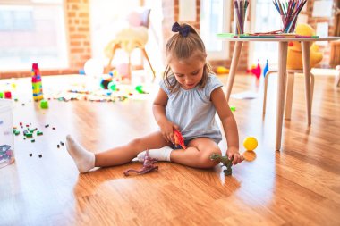 Genç, güzel sarışın kız anaokulunda oyuncaklarla oyun oynuyor, gülümseyerek evde dinozor oyuncaklarıyla oynuyor.