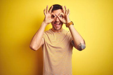 İzole edilmiş sarı arka plan üzerinde çizgili tişört giyen genç yakışıklı bir adam dürbün gibi jest yapıyor, gözleri parmaklarından bakıyor. Çılgın ifade.