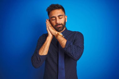 Genç Hintli iş adamı zarif gömlek ve kravat giyer. İzole edilmiş mavi arka plan üzerinde durur. Yorgun uyur, hayal kurar ve elleri ile birlikte poz verir..