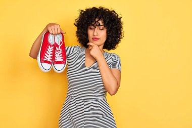 Kıvırcık saçlı genç bir Arap kadın izole edilmiş sarı arka planda spor ayakkabıları tutuyor soru soran ciddi bir yüz, çok karışık bir fikir.