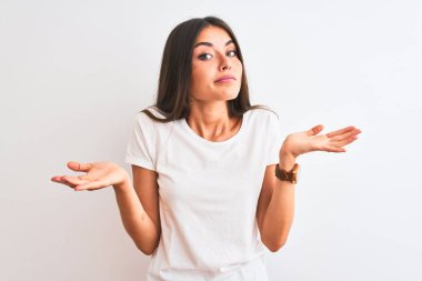 Genç güzel kadın rahat t-shirt izole beyaz arka plan üzerinde duran giyen habersiz ve kolları ve elleri ile karışık ifade kaldırdı. Şüphe kavramı.