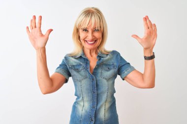 İzole edilmiş beyaz arka plan üzerinde duran sıradan bir kot gömlek giyen orta yaşlı kadın on numaralı parmaklarıyla işaret ederken kendinden emin ve mutlu bir şekilde gülümsüyor..