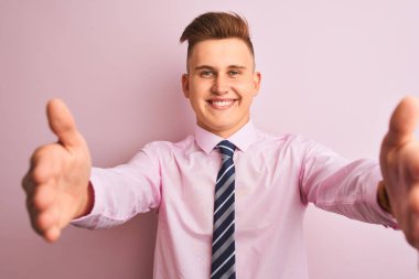 Gömlek ve kravat takan genç yakışıklı iş adamı pembe arka planda dikilip kameraya sarılmak için gülümseyerek bakıyor. Mutluluğu kucaklayan neşeli bir ifade..