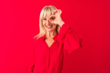 Orta yaşlı, zarif gömlekli bir kadın izole edilmiş kırmızı arka planda dikiliyor. El hareketleriyle gülümsüyor, gözleriyle mutlu yüzüyle parmaklarının arasından bakıyor..