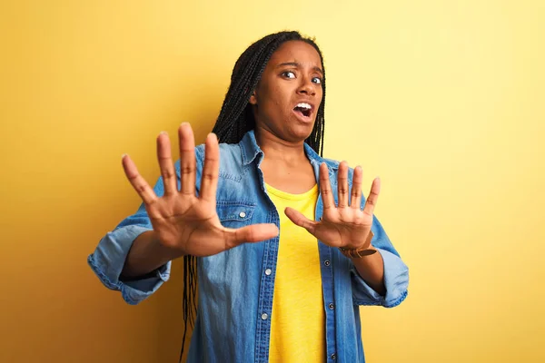 Young african american woman wearing denim shirt standing over isolated yellow background afraid and terrified with fear expression stop gesture with hands, shouting in shock. Panic concept.
