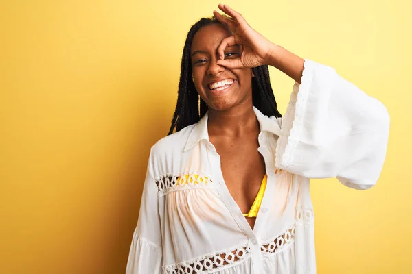 African american woman on vacation wearing bikini and shirt over isolated yellow background doing ok gesture with hand smiling, eye looking through fingers with happy face.