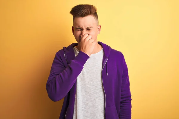 孤立した黄色の背景の上に立って紫色のスウェットシャツを着て若いハンサムな男は 鼻の上に指で息を保持し 臭いと嫌な 耐え難い匂いを嗅ぐ 悪い匂いの概念 — ストック写真