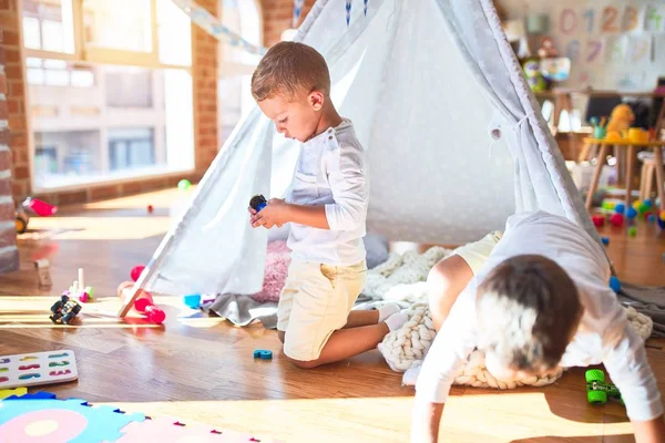 Adorable blonde twins playing inside tipi around lots of toys at kindergarten