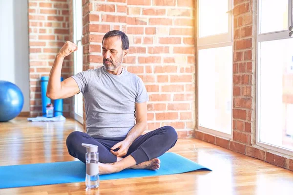 Middle age handsome sportman sitting on mat doing stretching yoga exercise at gym Strong person showing arm muscle, confident and proud of power