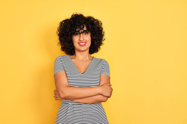 Молодая арабская женщина с вьющимися волосами в полосатом платье на изолированном желтом фоне счастливое лицо улыбается со скрещенными руками, глядя в камеру. Позитивный человек
.