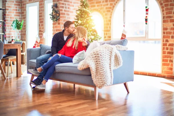 年轻漂亮的夫妇面带微笑 既快乐又自信 坐在沙发上拥抱和亲吻着家里的圣诞树 — 图库照片