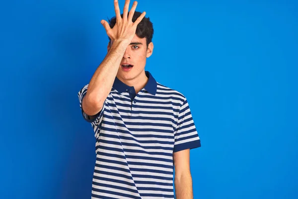 Teenager boy wearing casual t-shirt standing over blue isolated background surprised with hand on head for mistake, remember error. Forgot, bad memory concept.